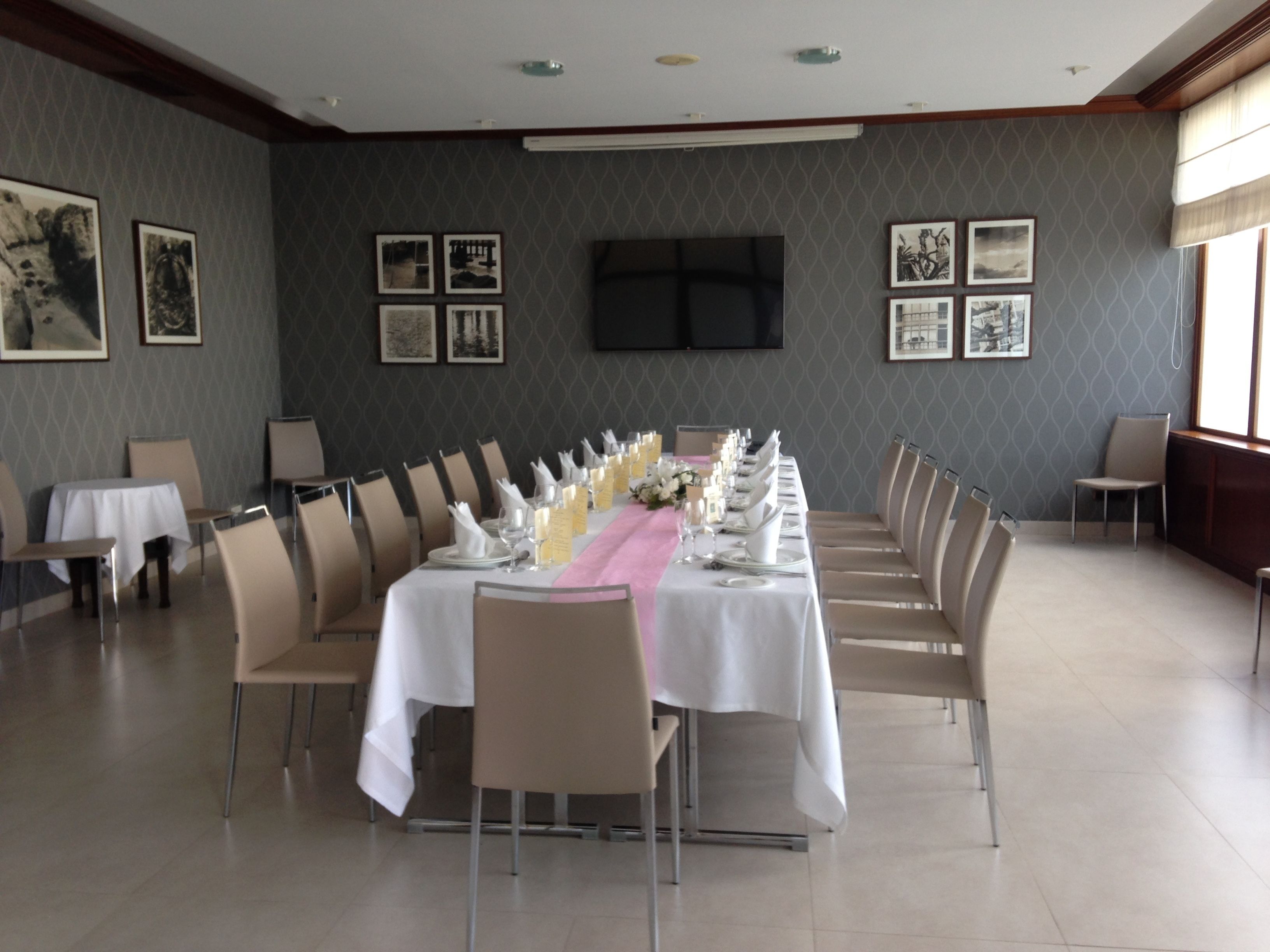 Un salón de conferencias con una larga mesa dispuesta para un evento formal, rodeado de sillas, con cuadros en las paredes y un televisor de pantalla plana montado sobre la mesa.
