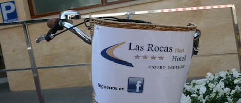 A wicker basket attached to a bicycle with a sign advertising "Las Rocas Playa Hotel CASTRO URDIALES" and a Facebook icon.