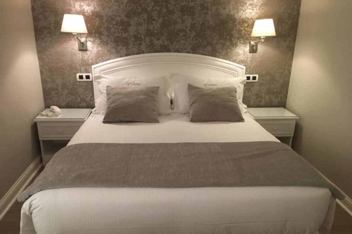 Una cama bien tendida con un cabecero blanco, dos lámparas de noche y una mesita de noche con un teléfono.
