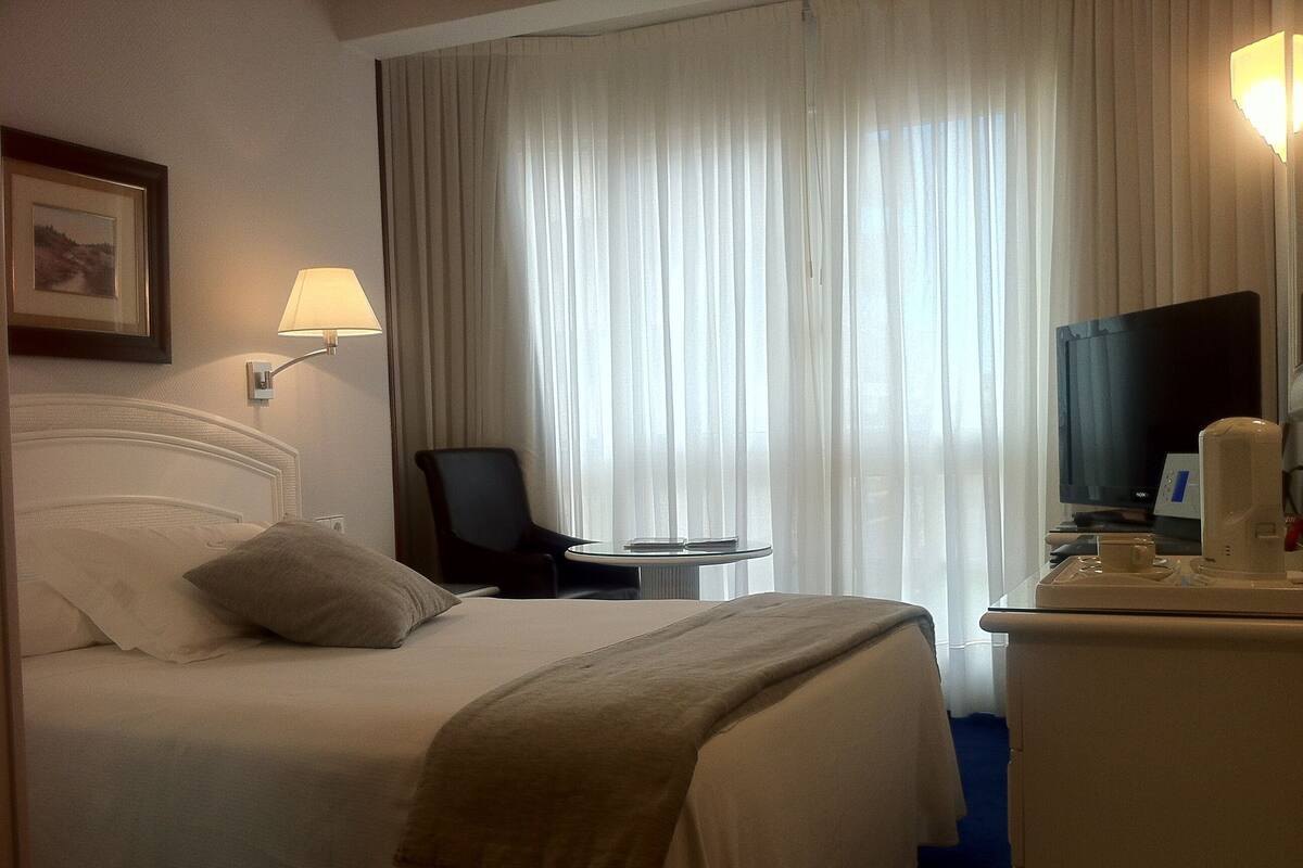Una habitación de hotel con una cama, una silla, una mesita, un televisor y una lámpara.