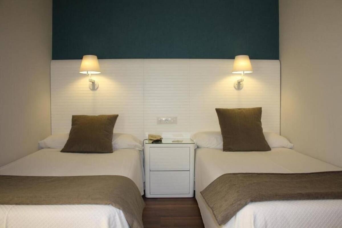 Habitación de hotel con dos camas, cada una con un almohadón marrón y una colcha blanca. Hay dos lámparas fijadas a la pared, una a cada lado de las camas.