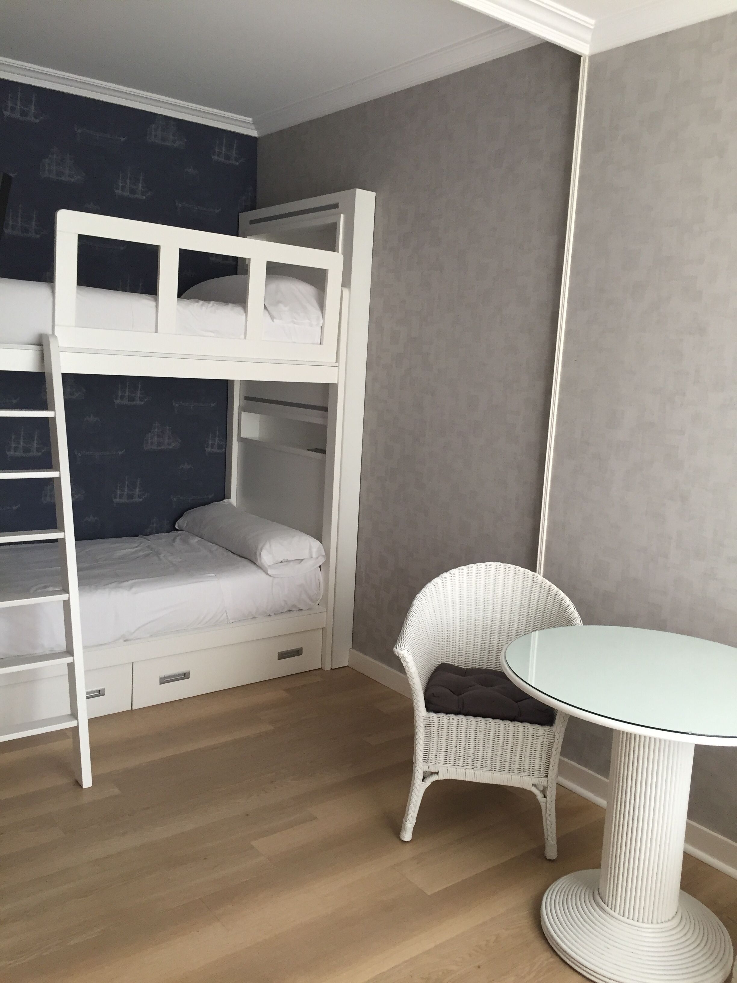 Ein Etagenbett mit unterem Bett, ein weißer Stuhl und ein kleiner runder Tisch in einem Zimmer mit Holzboden und grauen Wänden.