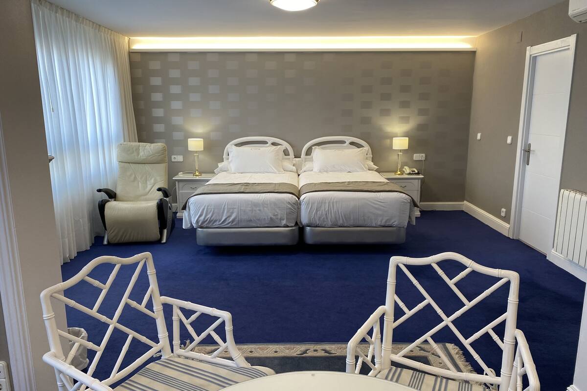 Habitación de hotel con dos camas, una silla, una mesa y vista a la calle.