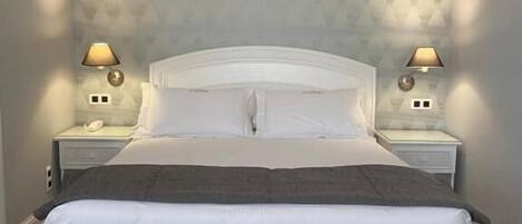 Una cama grande con cabecera gris, dos mesitas de noche con lámparas y papel tapiz estampado.