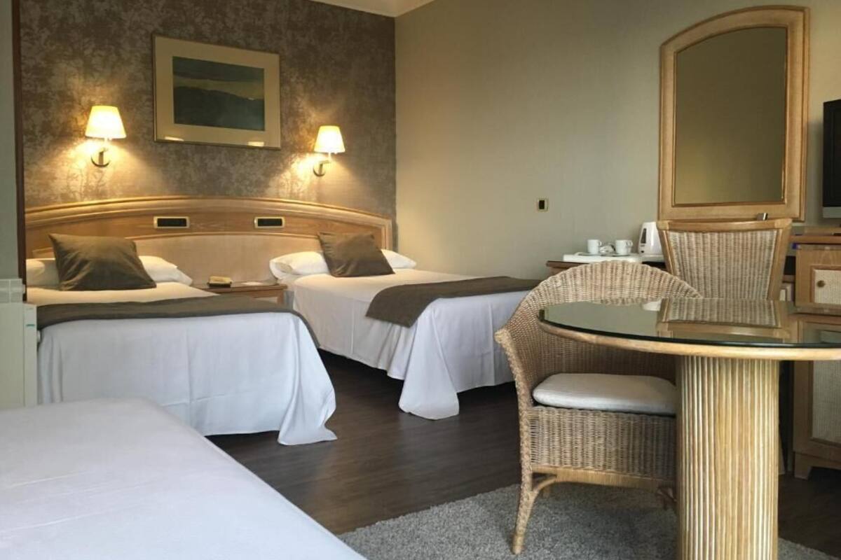 Habitación de hotel con dos camas, una mesa redonda con sillas, un espejo grande y lámparas fijadas a la pared.