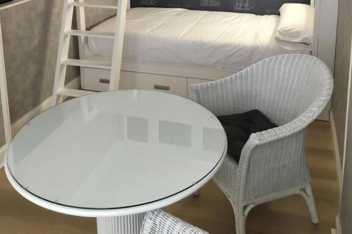Una cama tipo litera con escalera, una mesita redonda pequeña y una silla de mimbre en una habitación con papel tapiz estampado.
