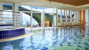 An indoor swimming pool with a wooden ceiling, large windows offering a view of a forested hillside, and a seating area with lounge chairs. - OREA Resort Sklář Harrachov (Harrachov)