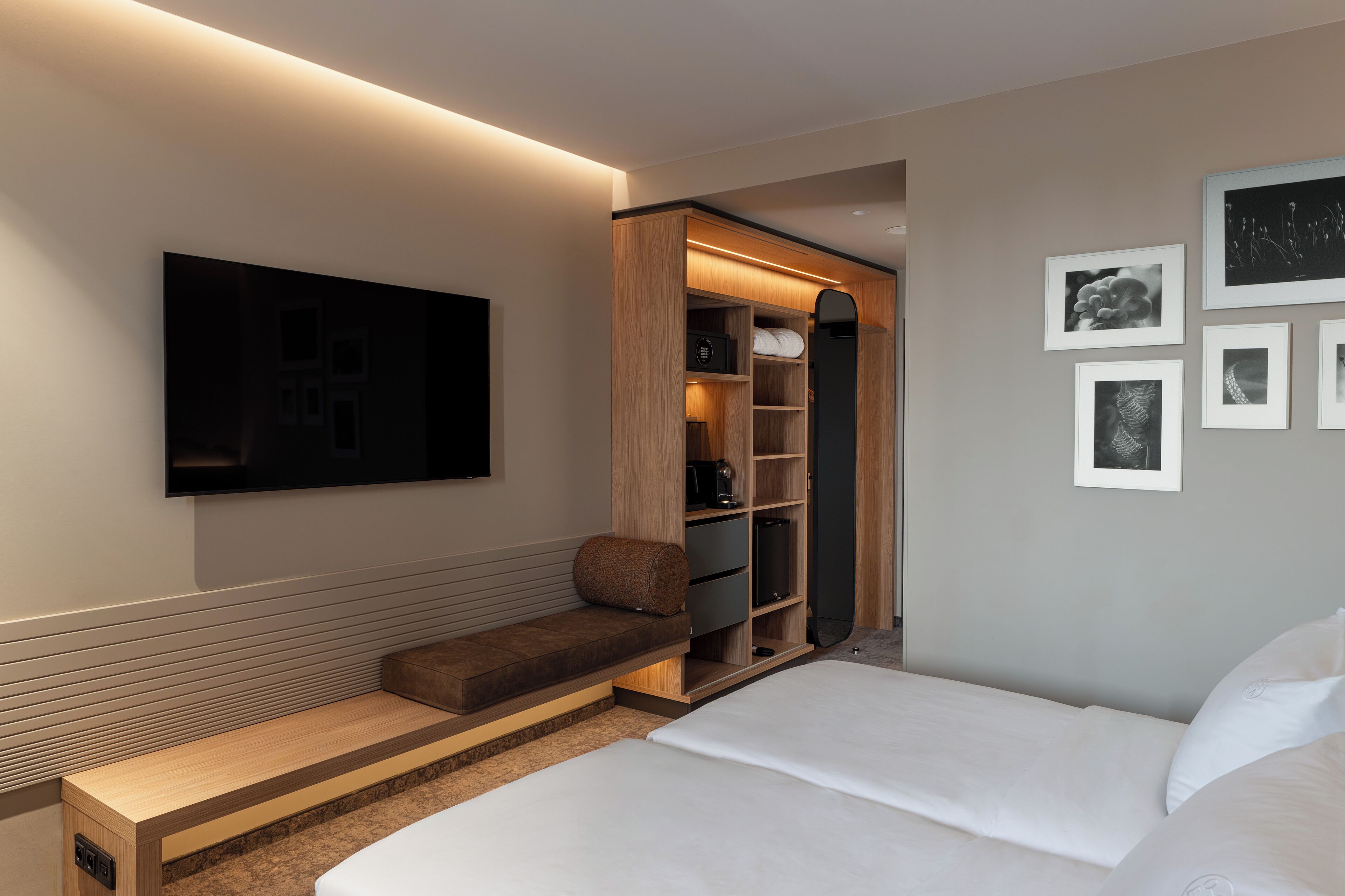 A modern hotel room with a bed, a bench, a walk-in closet, and a flat-screen TV mounted on the wall.