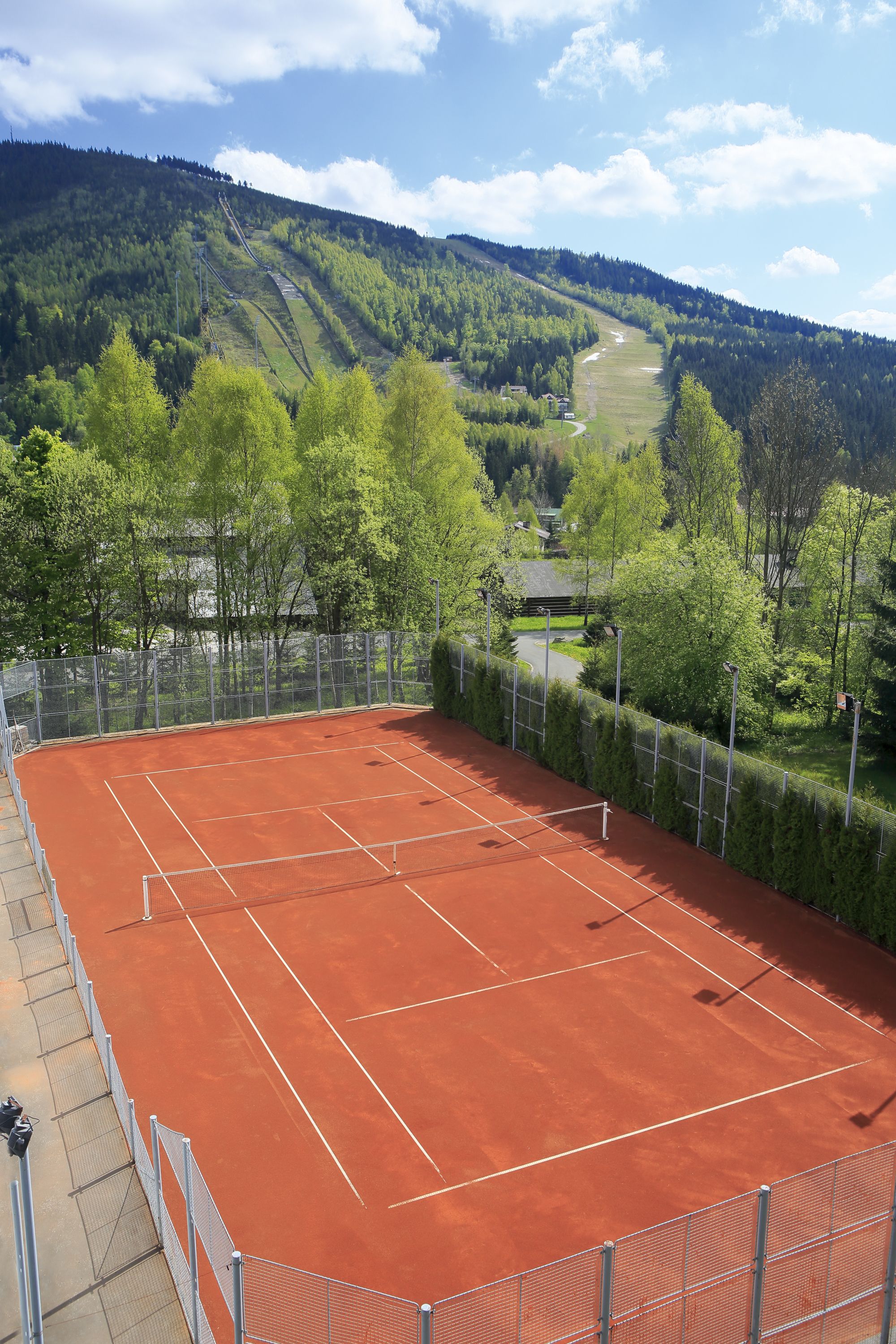 En tennisbane med hegn og træer, foran et bakket landskab.
