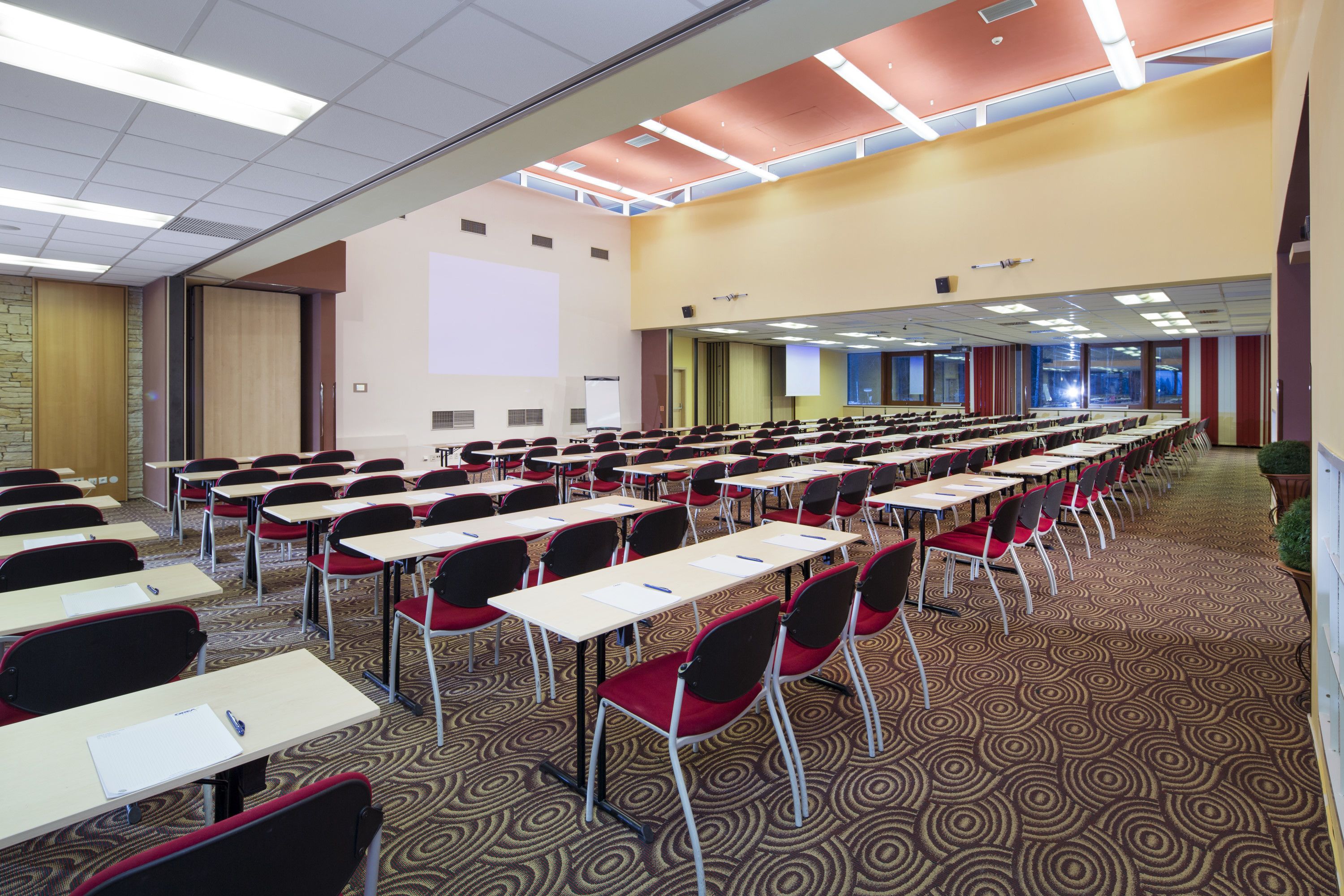 Une salle de conférence avec des rangées de tables et de chaises, un écran de projection et un sol recouvert de moquette.
