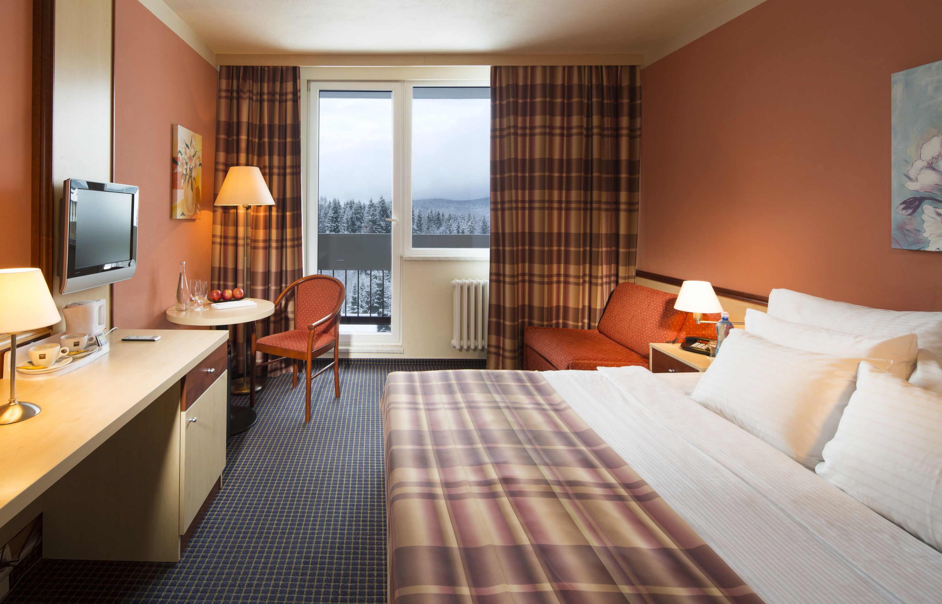 A hotel room with a bed, a desk, a chair, a TV, and a window with a view of trees.