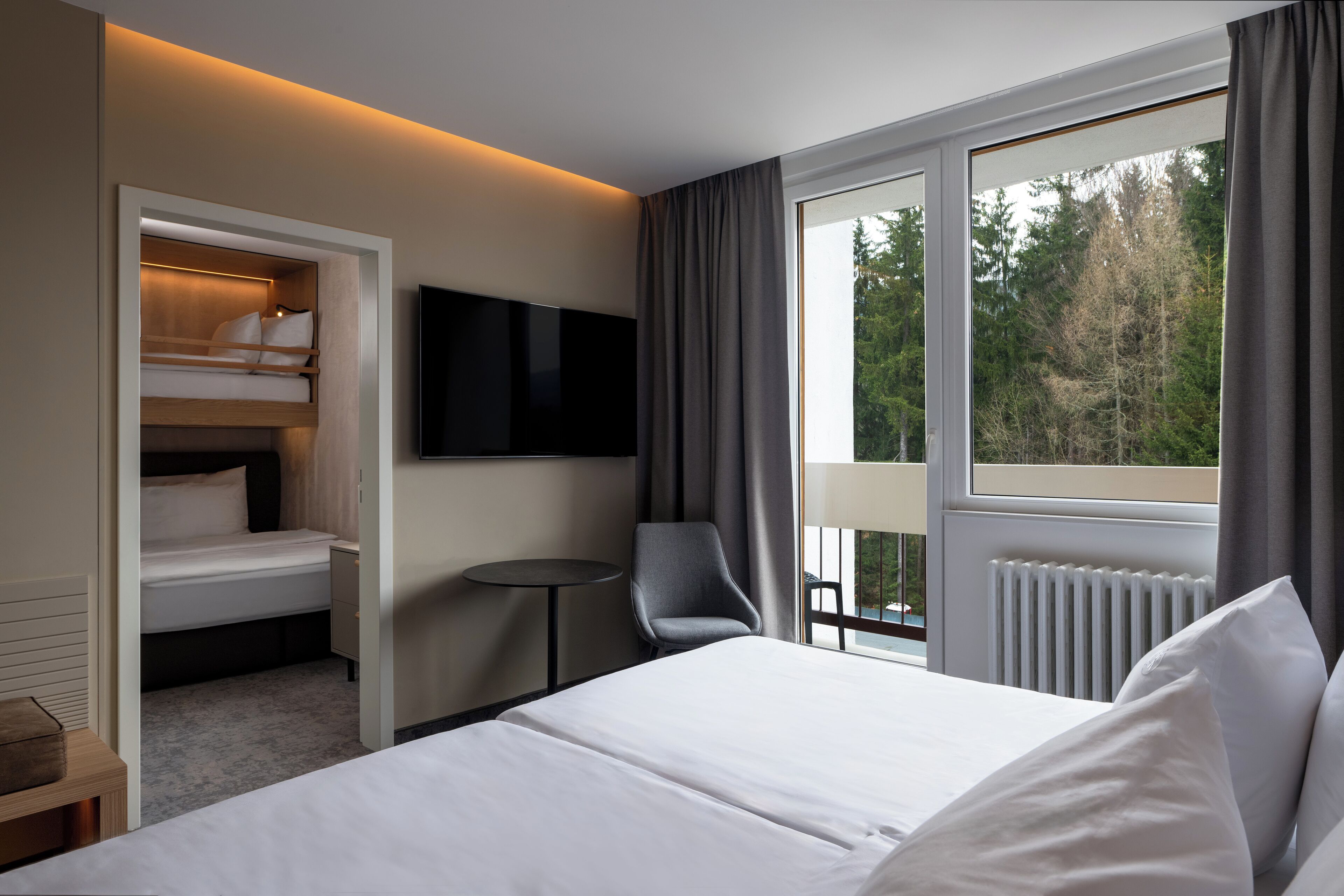 A hotel room with a bed, a TV, a chair, a small table, and a window with a view of trees.