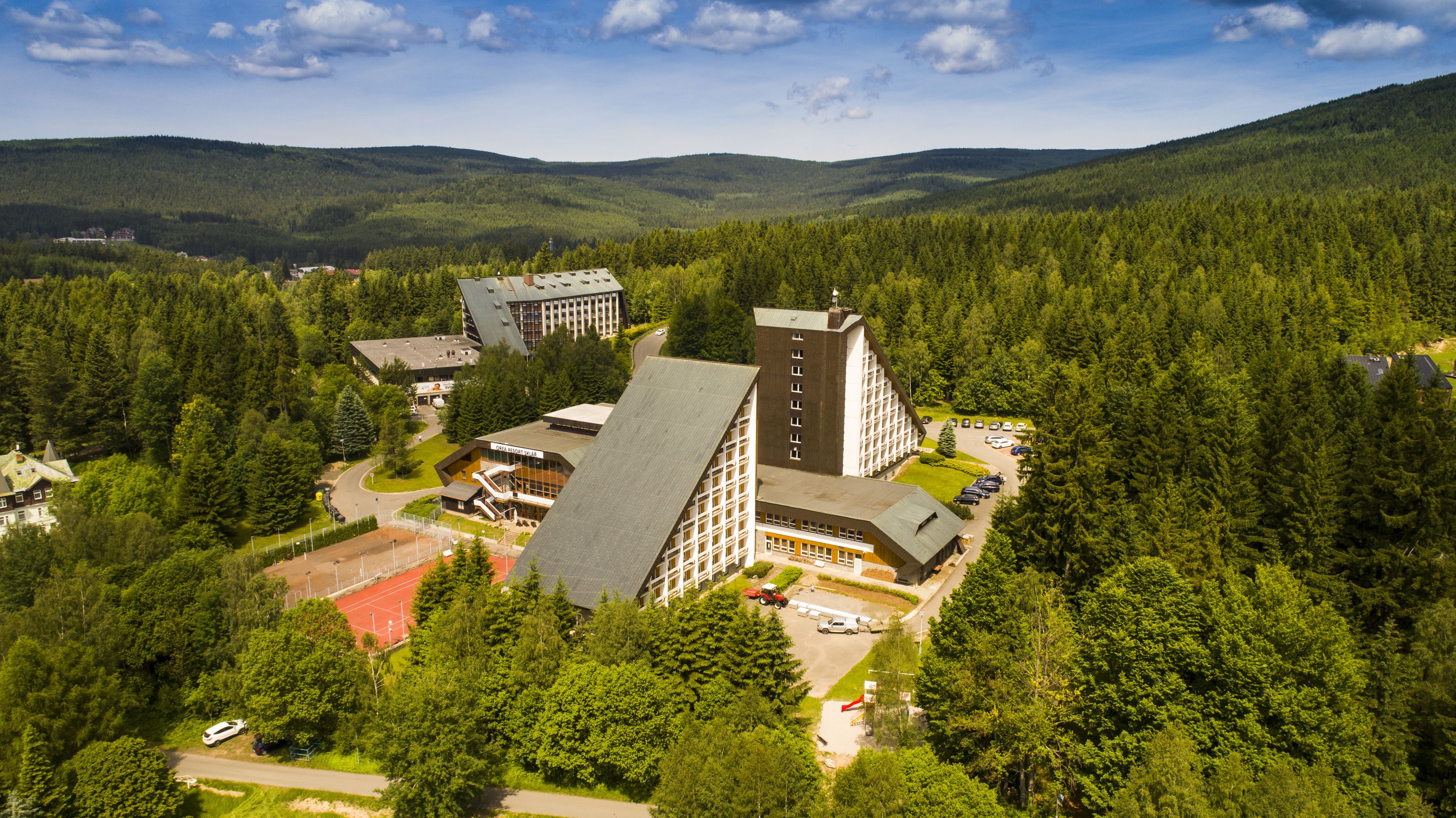Vista aérea de un resort con varias edificaciones, incluyendo un hotel grande con un techo triangular característico, rodeado de un bosque denso y colinas al fondo.