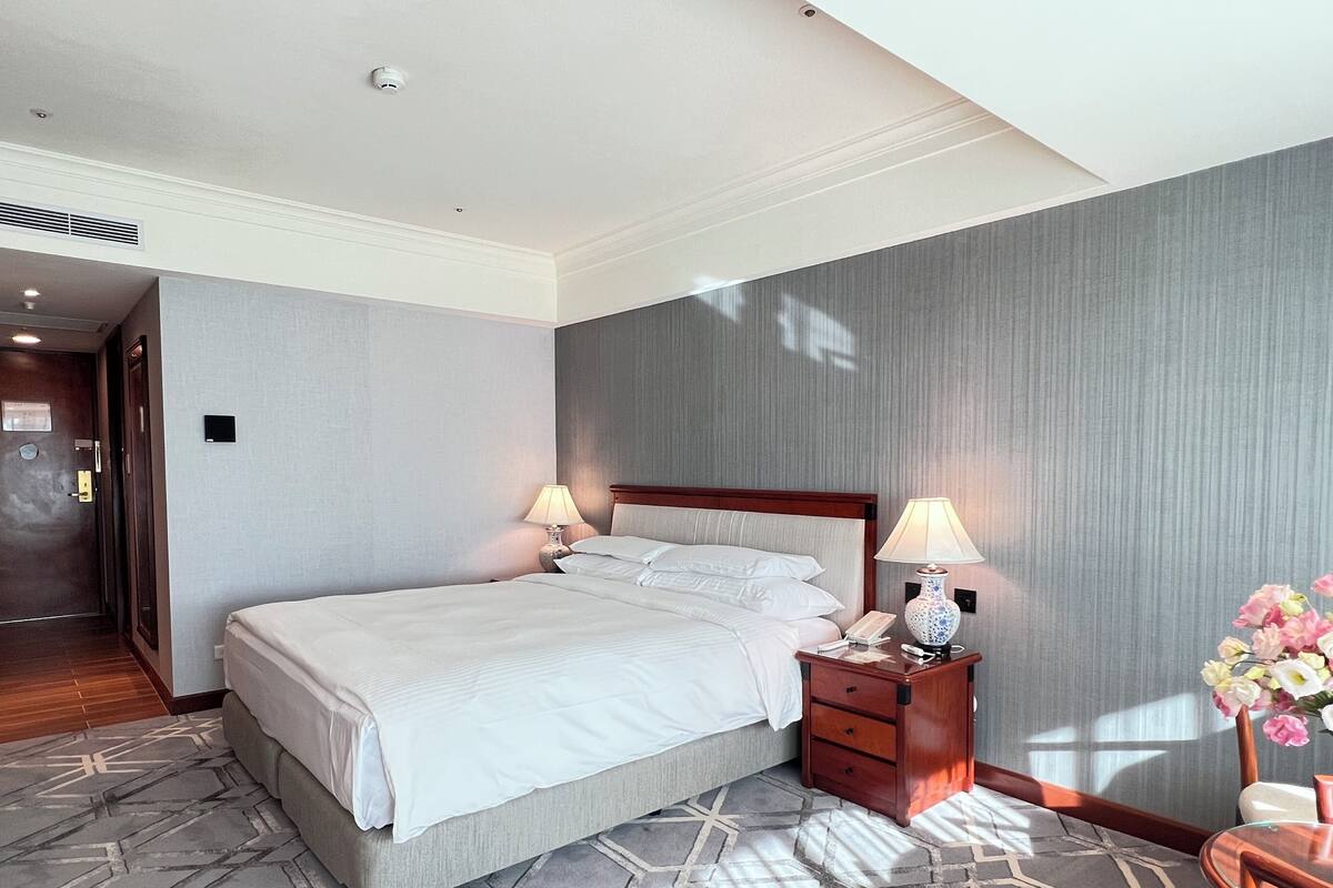 Une chambre d’hôtel avec un grand lit, deux tables de chevet avec des lampes, une petite table en bois et un vase de fleurs.