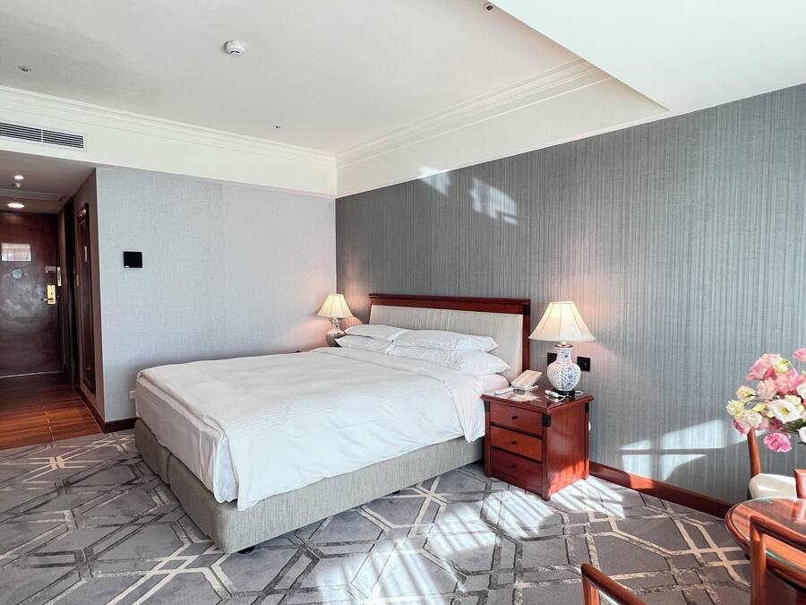 A hotel room with a large bed, two bedside tables with lamps, a wooden nightstand, and a vase of flowers.