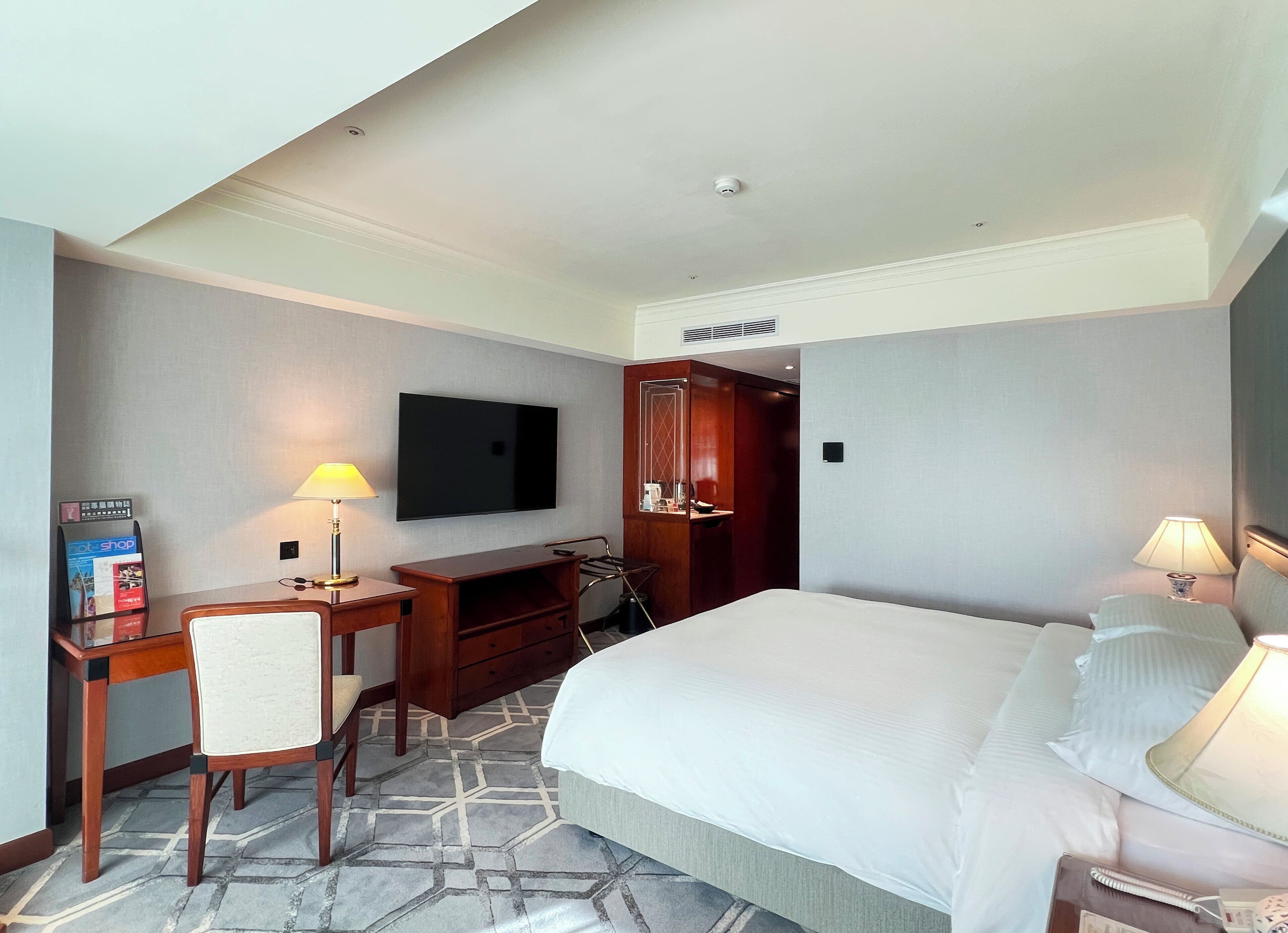A hotel room with a large bed, a desk with a chair, a television mounted on the wall, and a small table with a lamp.