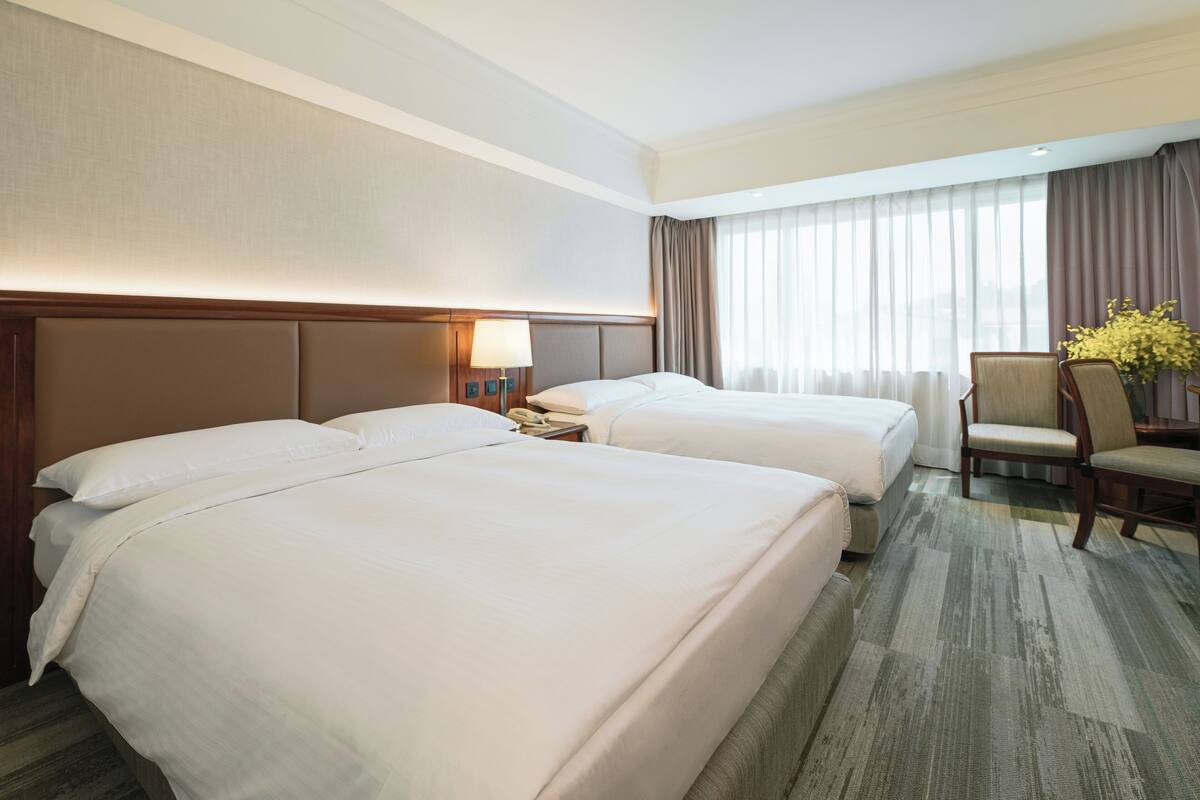 Une chambre d’hôtel avec deux lits, un bureau, une chaise et une grande fenêtre avec des rideaux.