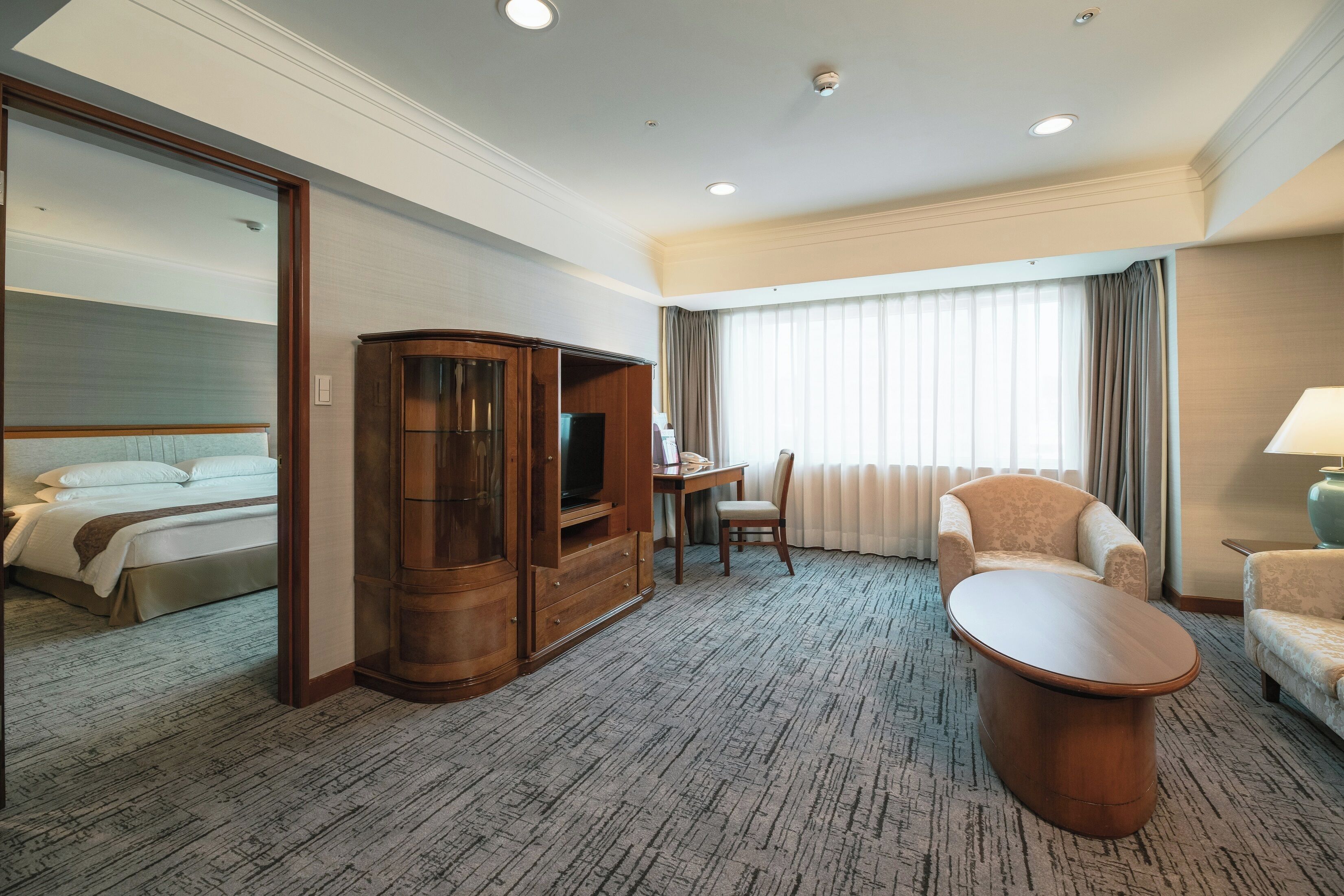 A hotel room with a bed, a wooden cabinet, a desk with a chair, a round coffee table, and a window with curtains.
