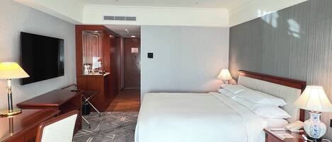 Una habitación de hotel con una cama grande, un escritorio con televisor de pantalla plana, una silla, una mesita de noche con lámpara y una alfombra estampada.