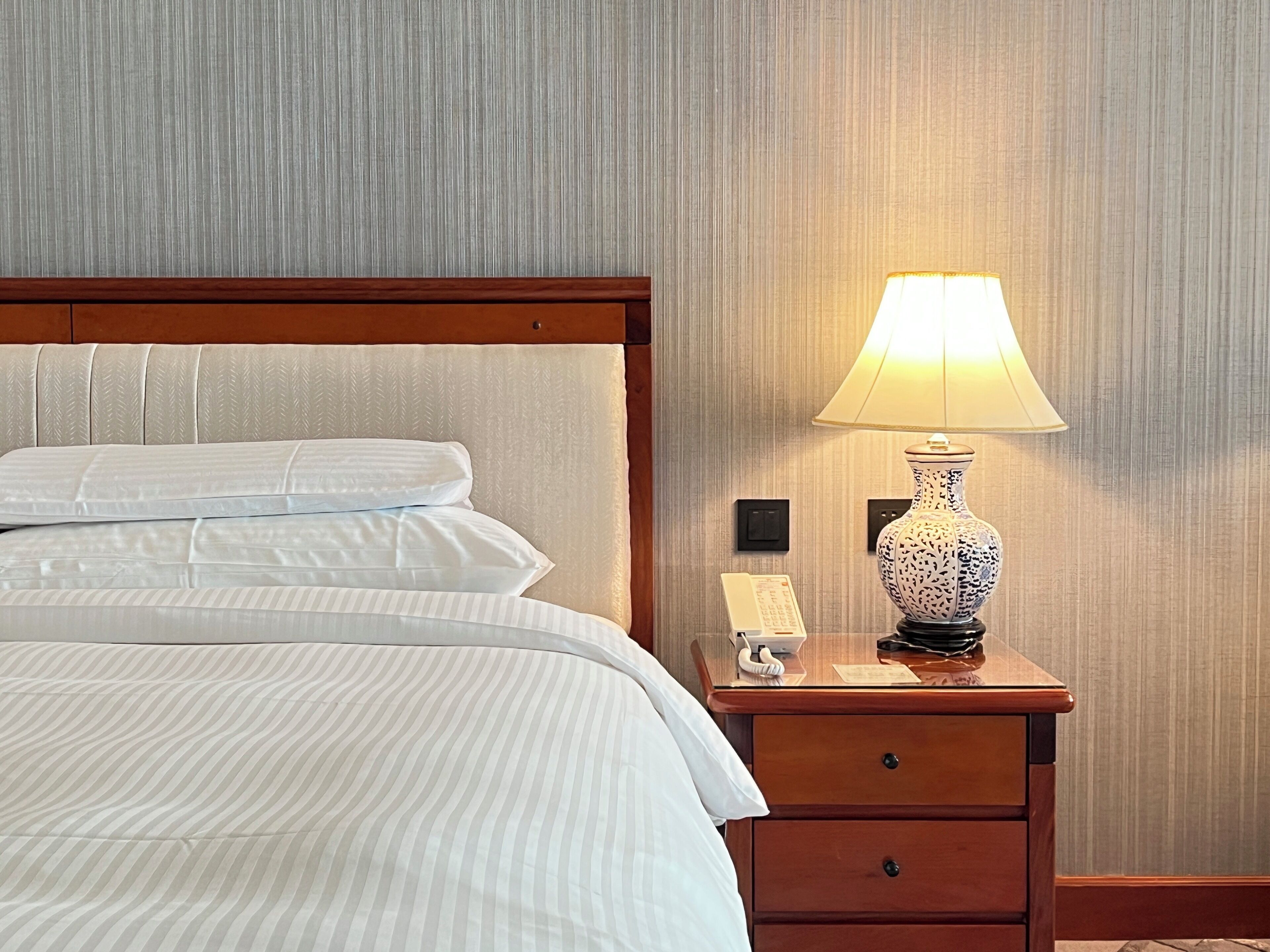 Una cama bien tendida con sábanas blancas, una mesita de noche de madera con una lámpara y un teléfono.