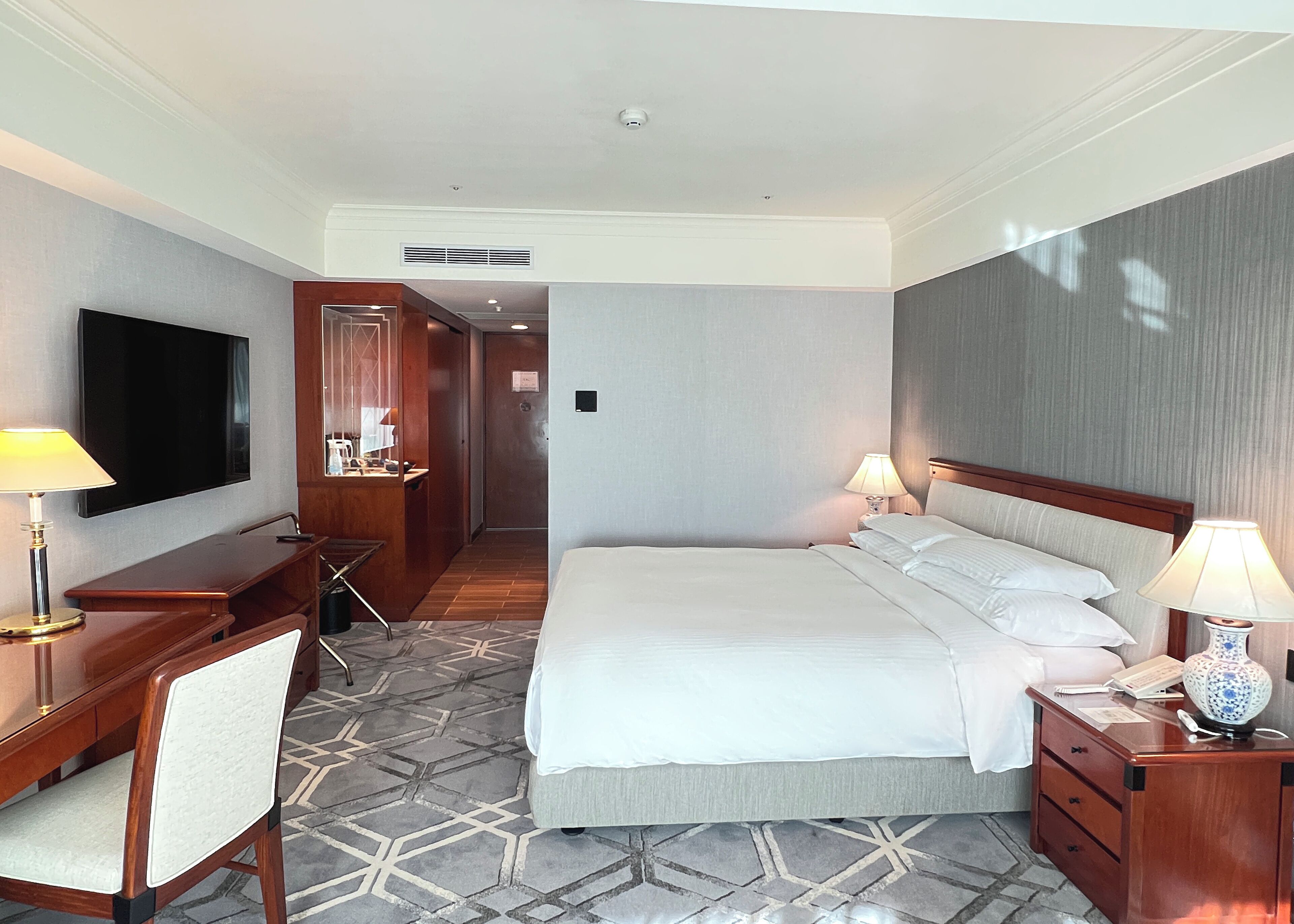 A hotel room with a large bed, a desk with a flat-screen TV, a chair, a bedside table with a lamp, and a patterned rug.