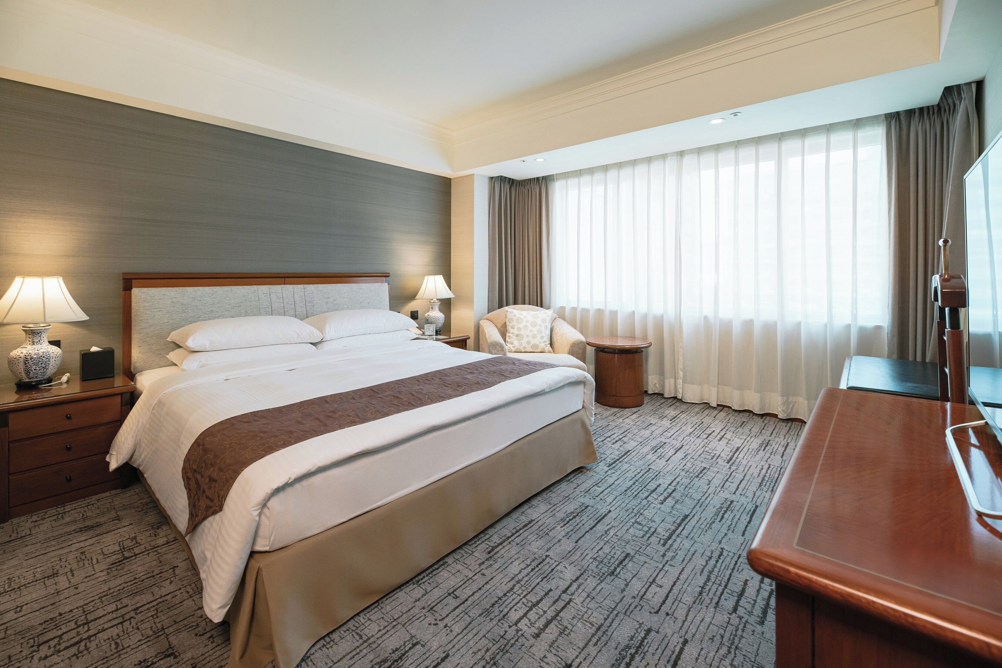 Ein Hotelzimmer mit einem großen Bett, zwei Nachttischen mit Lampen, einem Sessel, einem kleinen runden Tisch und einem Fernseher.
