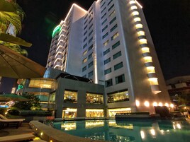 Un hotel con área de piscina, varios pisos y ventanas iluminadas.