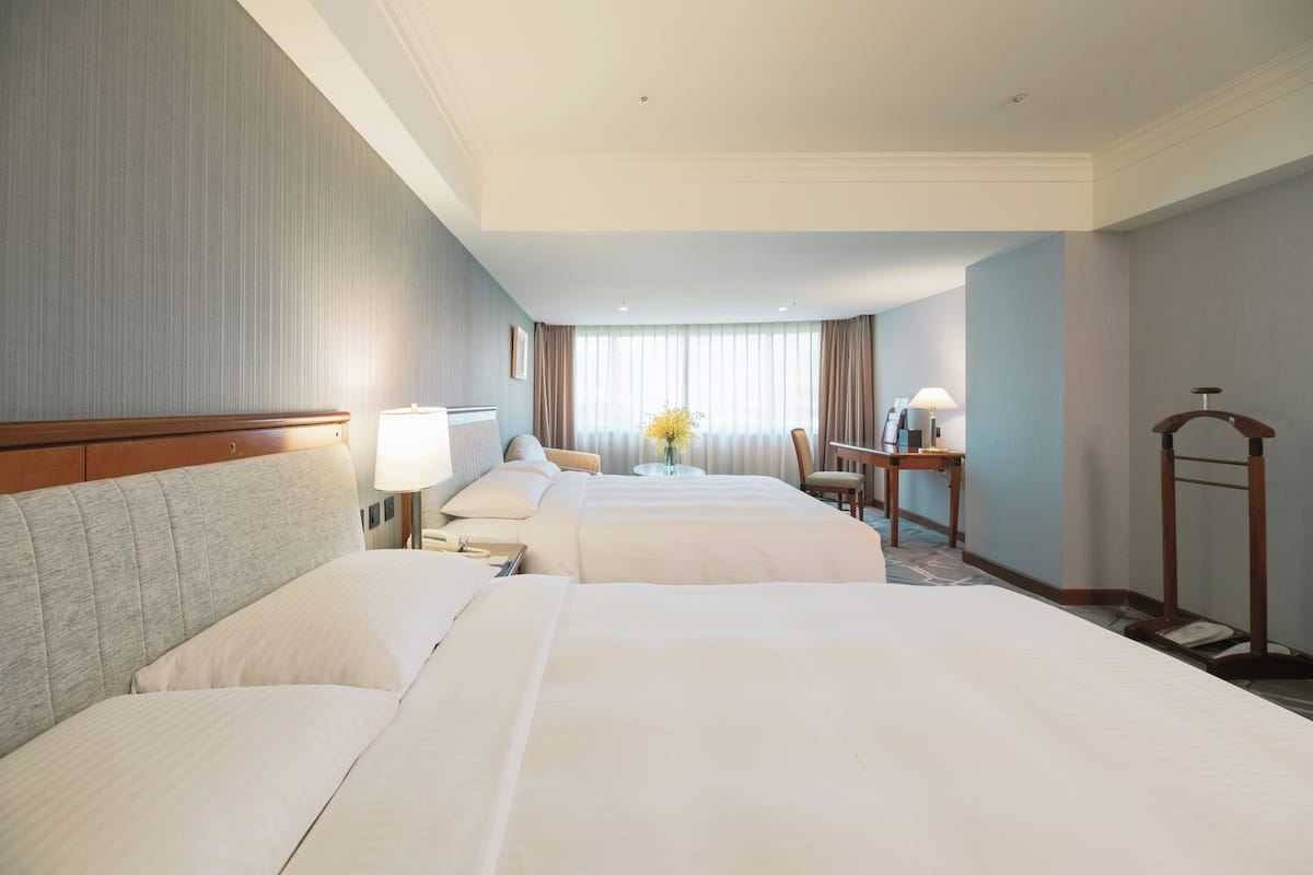 Une chambre d’hôtel avec deux lits, un bureau, une chaise, une lampe et un vase avec des fleurs.