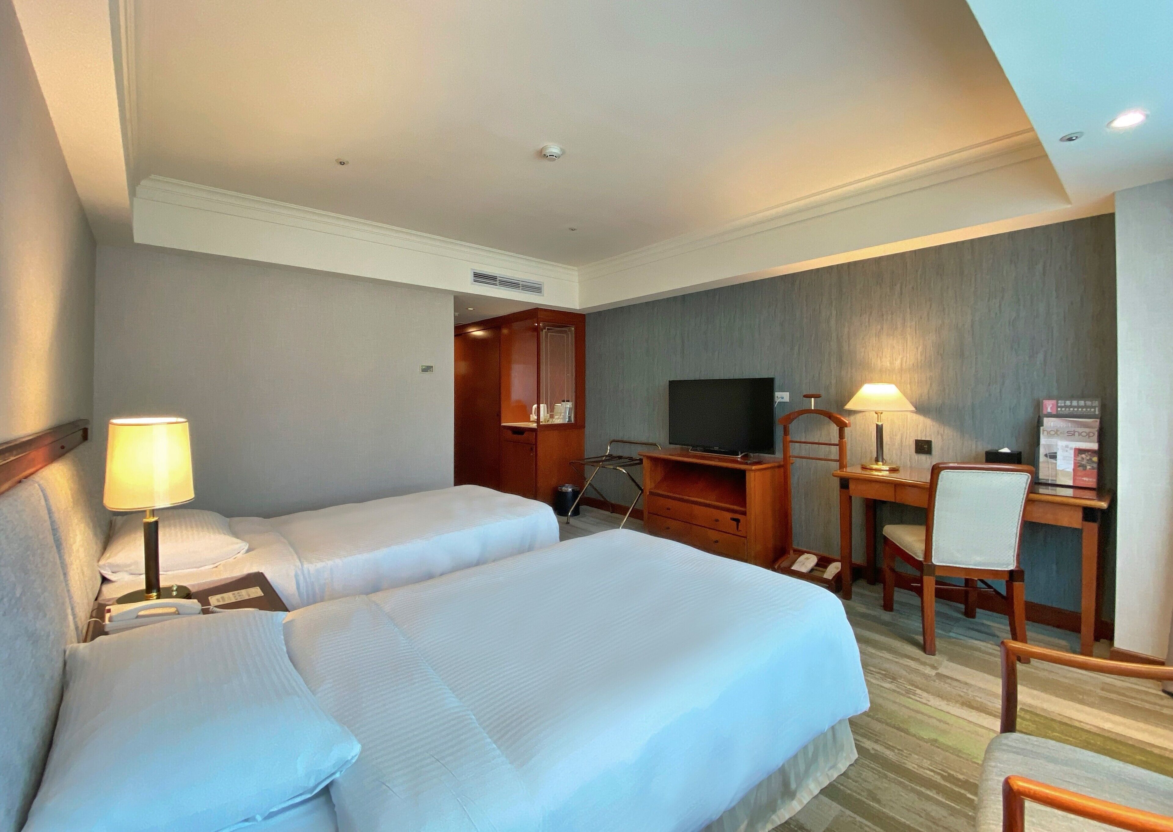 Habitación de hotel con dos camas, un escritorio con televisor, una silla y una lámpara.