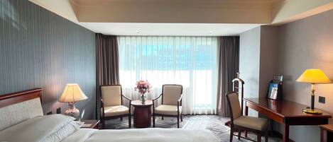 A hotel room with a large bed, two armchairs, a small table with a vase, a desk with a lamp, and a window with curtains.