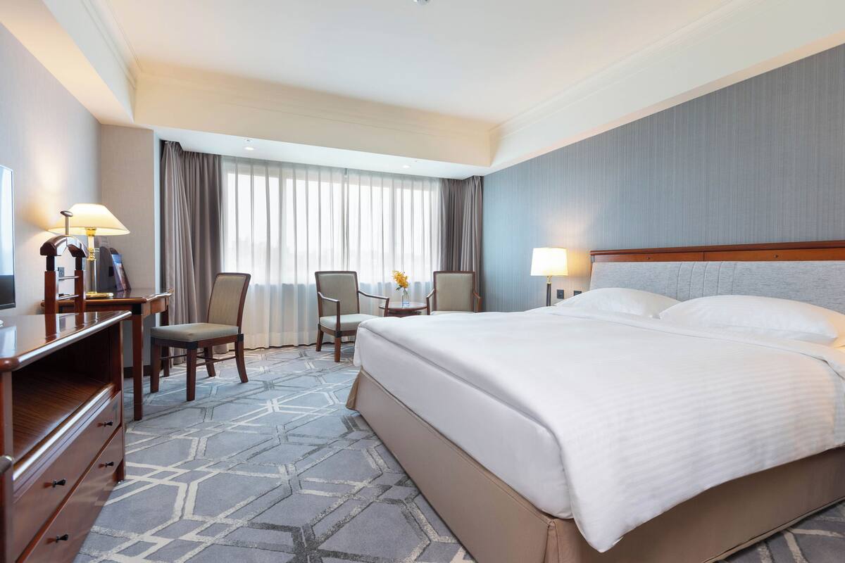 Une chambre d’hôtel avec un grand lit, un bureau avec une lampe, une chaise et une fenêtre avec des rideaux.