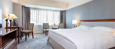 A hotel room with a large bed, a desk with a lamp, a chair, and a window with curtains.