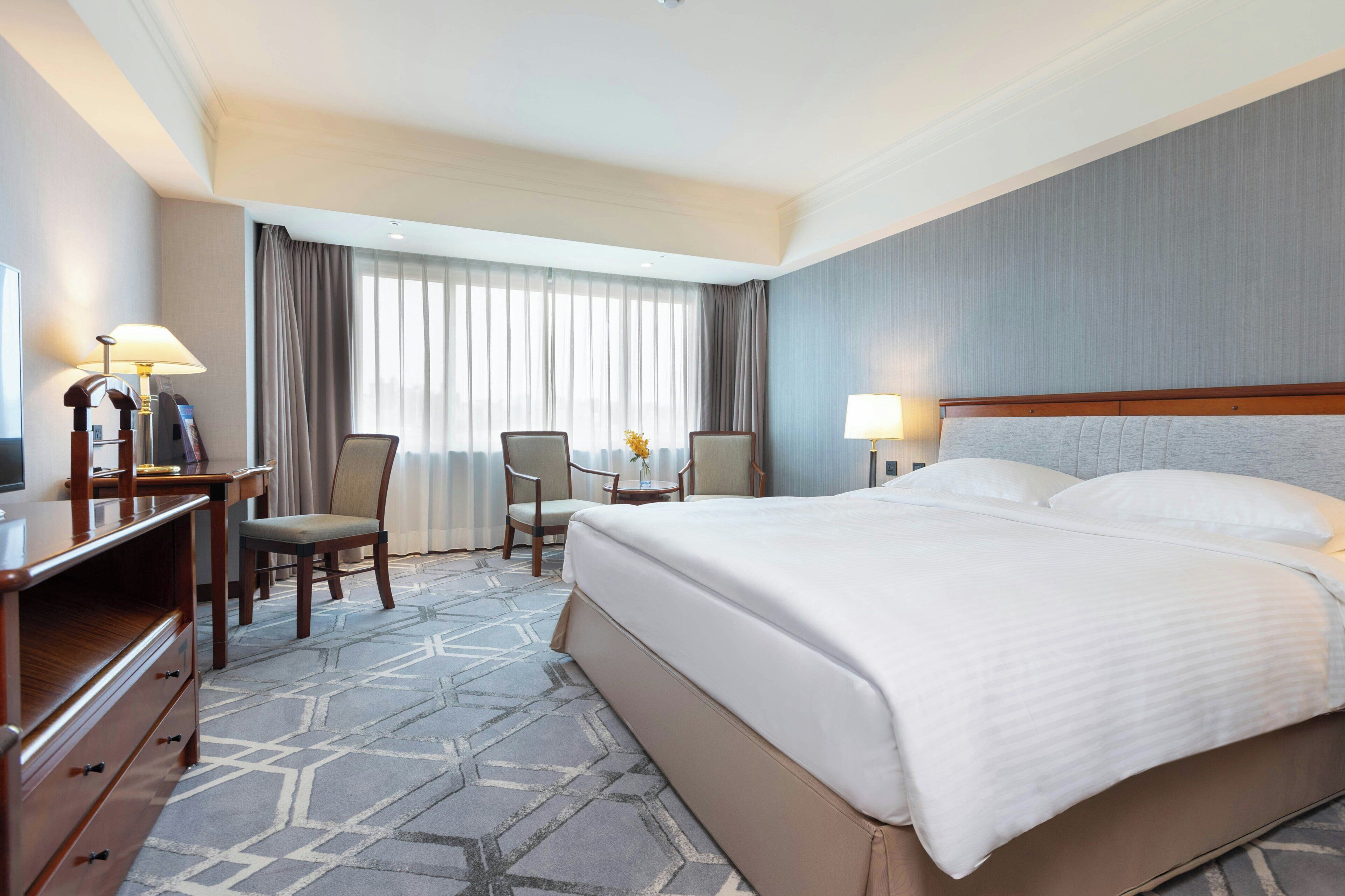 A hotel room with a large bed, a desk with a lamp, two chairs, and a view of the outdoors through the window.