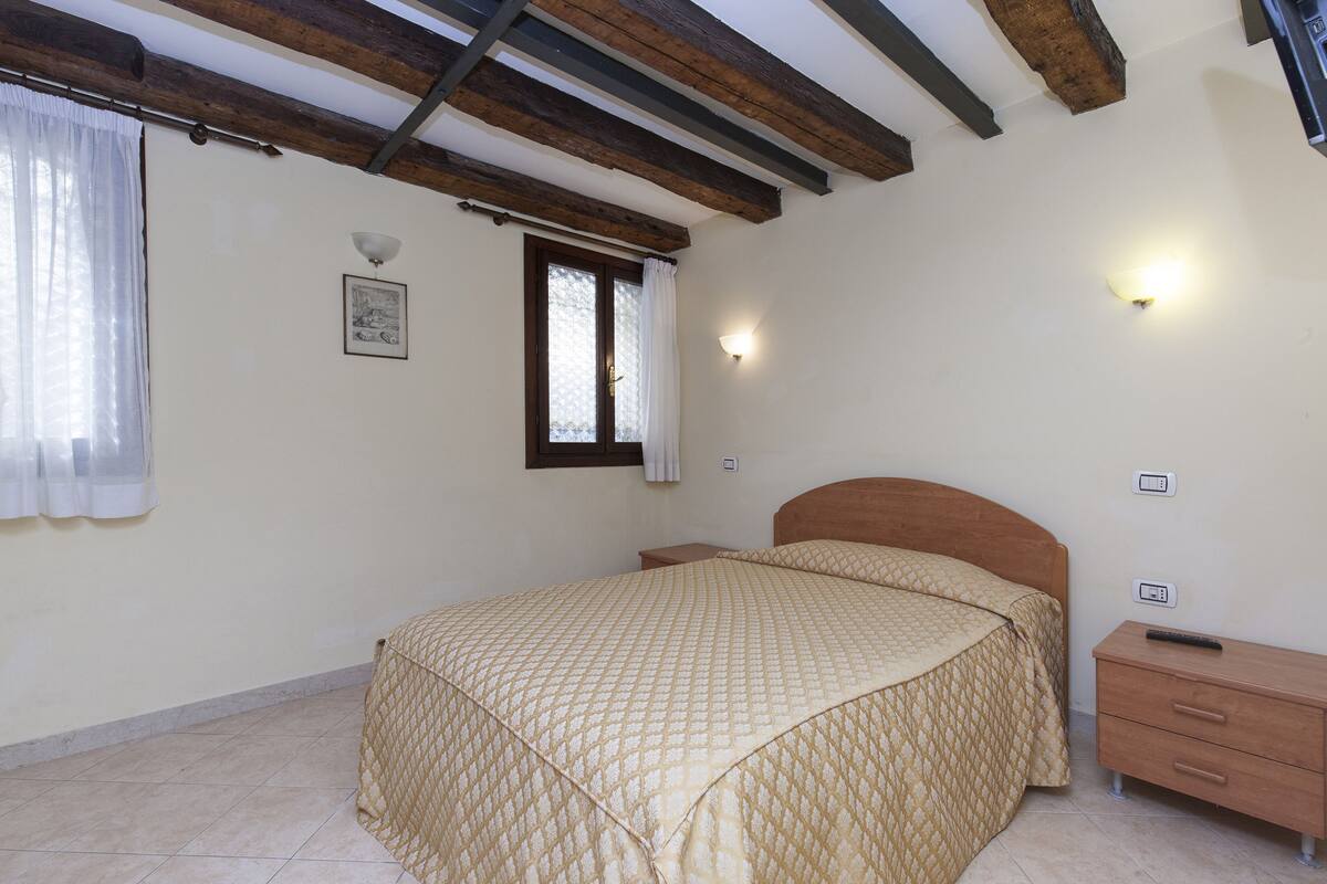 A bedroom with a bed, a nightstand, a wall-mounted light, a window with curtains, and a ceiling with exposed beams.