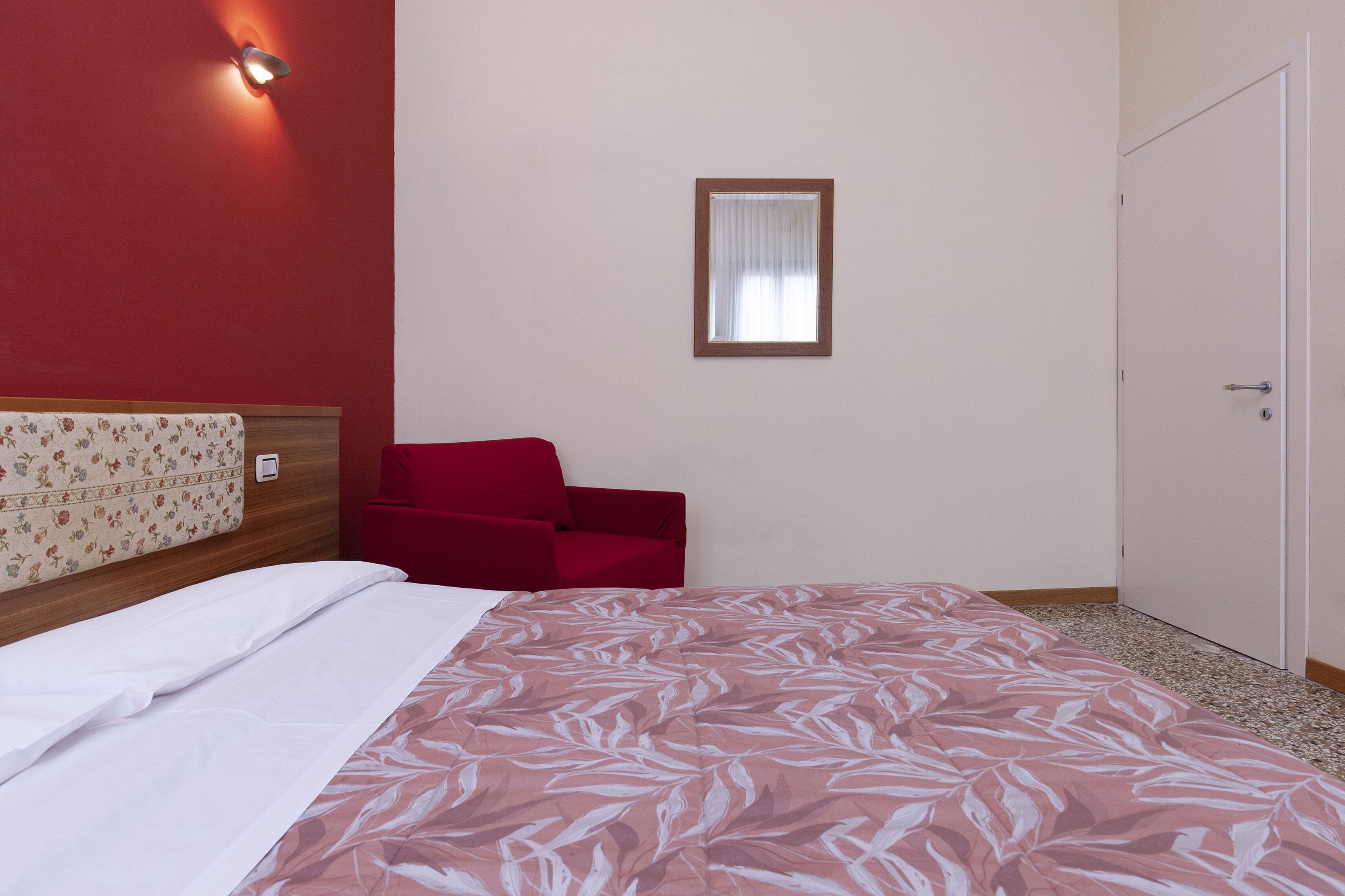 A hotel room with a bed, a red chair, a wall-mounted light, and a door.