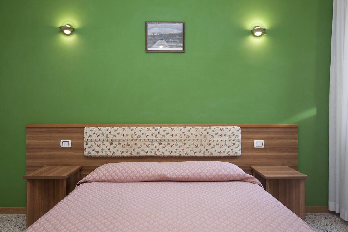 A bedroom with a green wall, a wooden headboard with a floral pattern, two bedside tables, a bed with a pink quilt, and two wall-mounted lights.