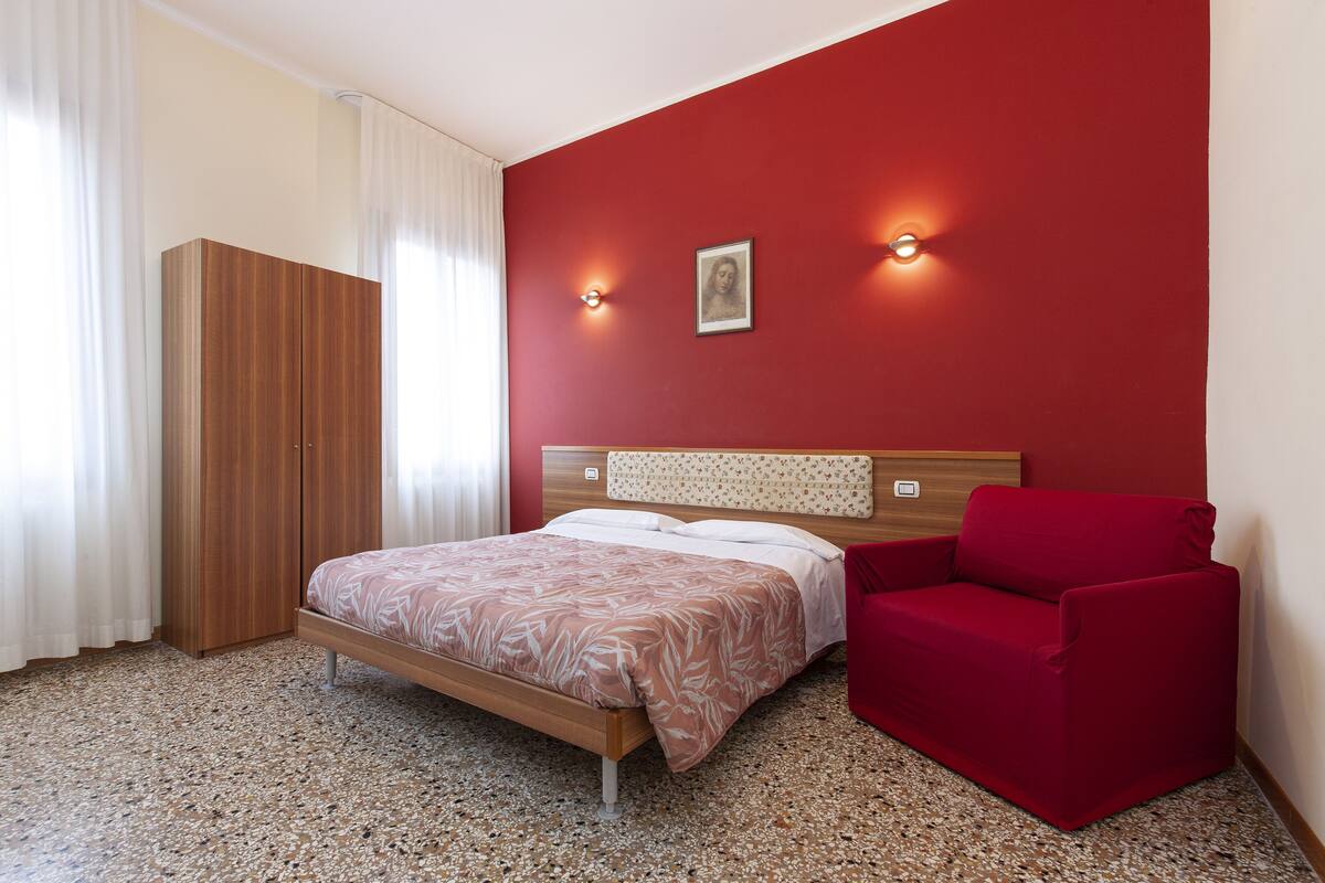 A bedroom with a bed, a red armchair, a wardrobe, and a wall-mounted picture.