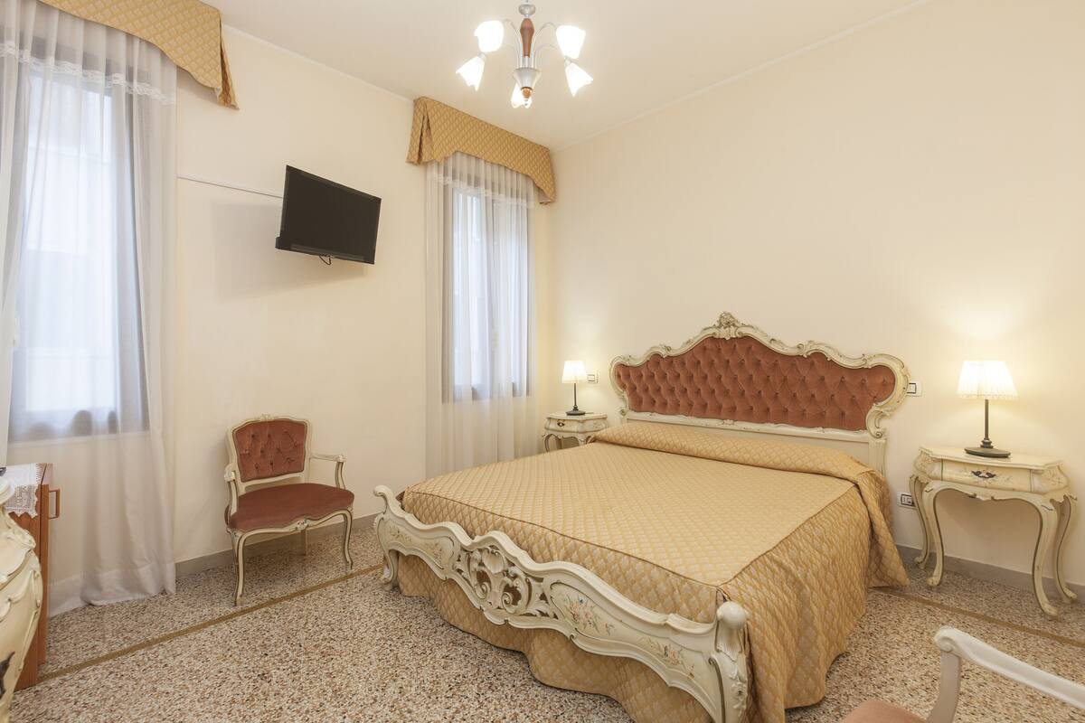 A bedroom with a large bed, two bedside tables with lamps, a chair, a television on the wall, and a window with sheer curtains.