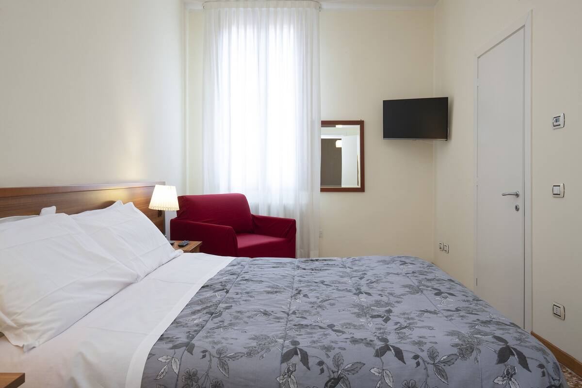 A hotel room with a bed, a red chair, a TV, and a door.
