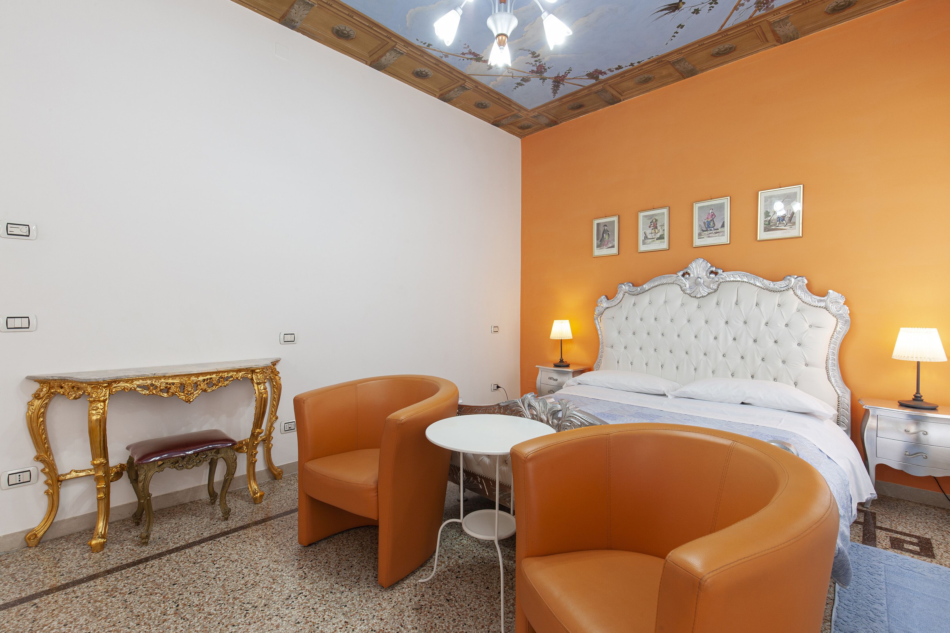 A hotel room with a bed, two orange chairs, a white table, and a gold-framed console table.