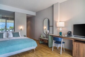 A modern hotel room with a large bed, a desk with a chair, a TV, and a window with a view of mountains. - Hotel Val di Sogno (Malcesine)