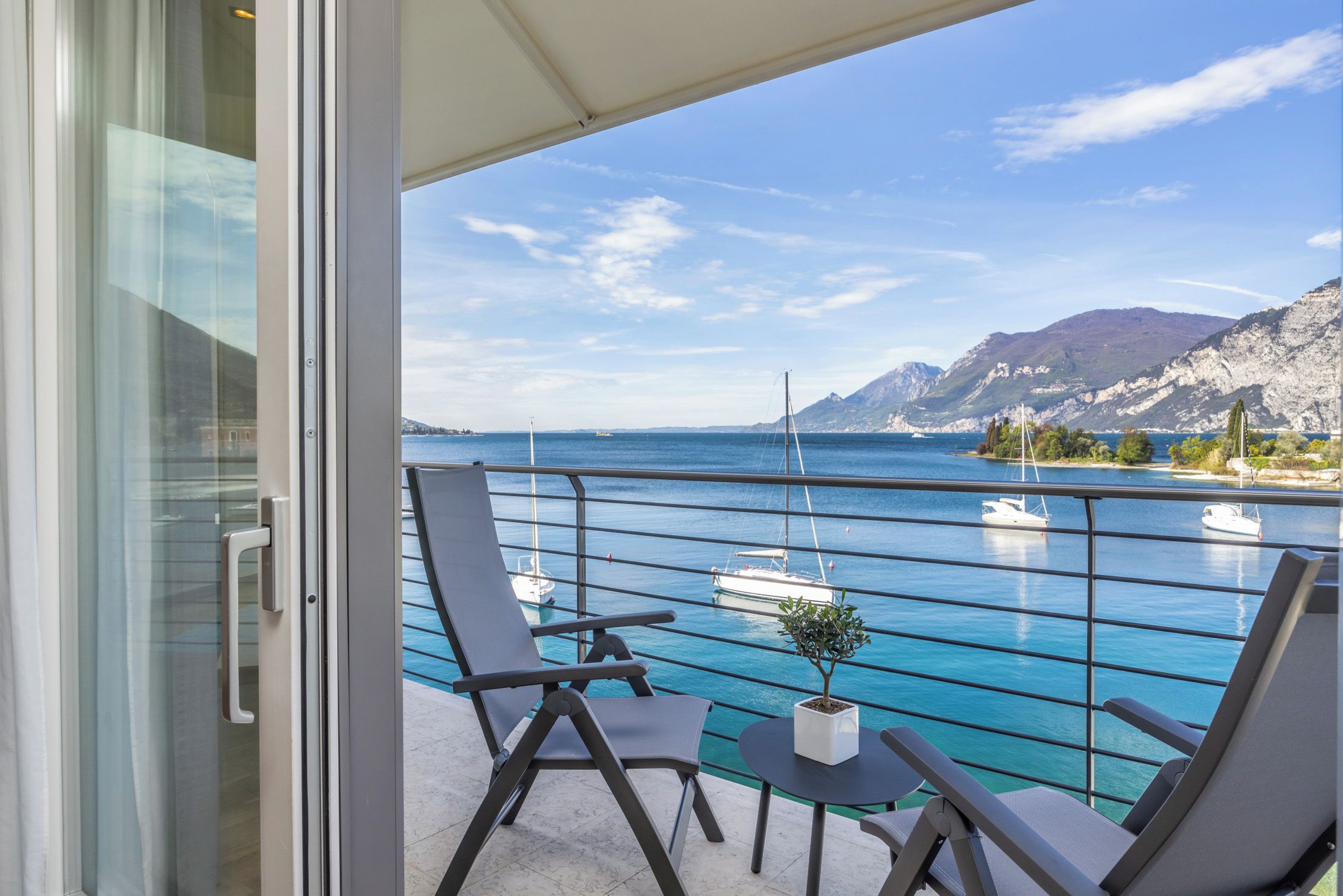 A balcony with two chairs and a small table, overlooking a marina with boats and a mountainous coastline.