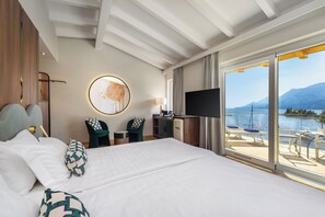 Habitación de hotel con una cama grande, un escritorio con televisor, una silla, una mesita y vistas a un lago y montañas.