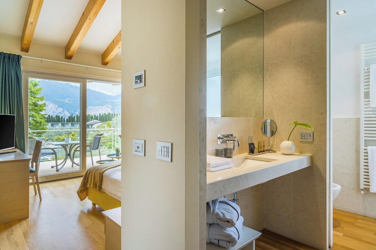 A modern hotel room with a balcony, a large mirror, a sink, and a view of mountains.