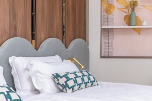 A neatly made bed with white linens and a patterned pillow, a bedside lamp, and a wooden wardrobe in the background.