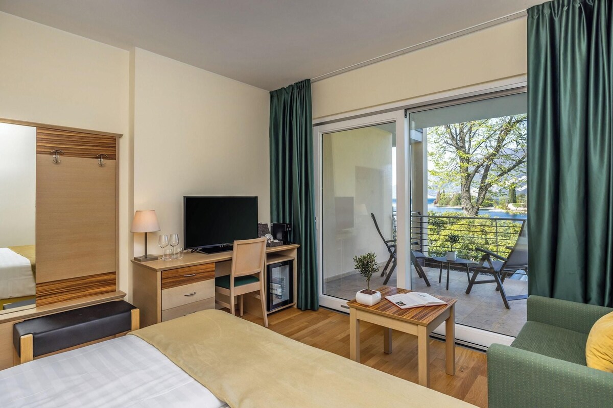 Habitación de hotel con cama, escritorio, silla, televisión y balcón con vista a árboles.