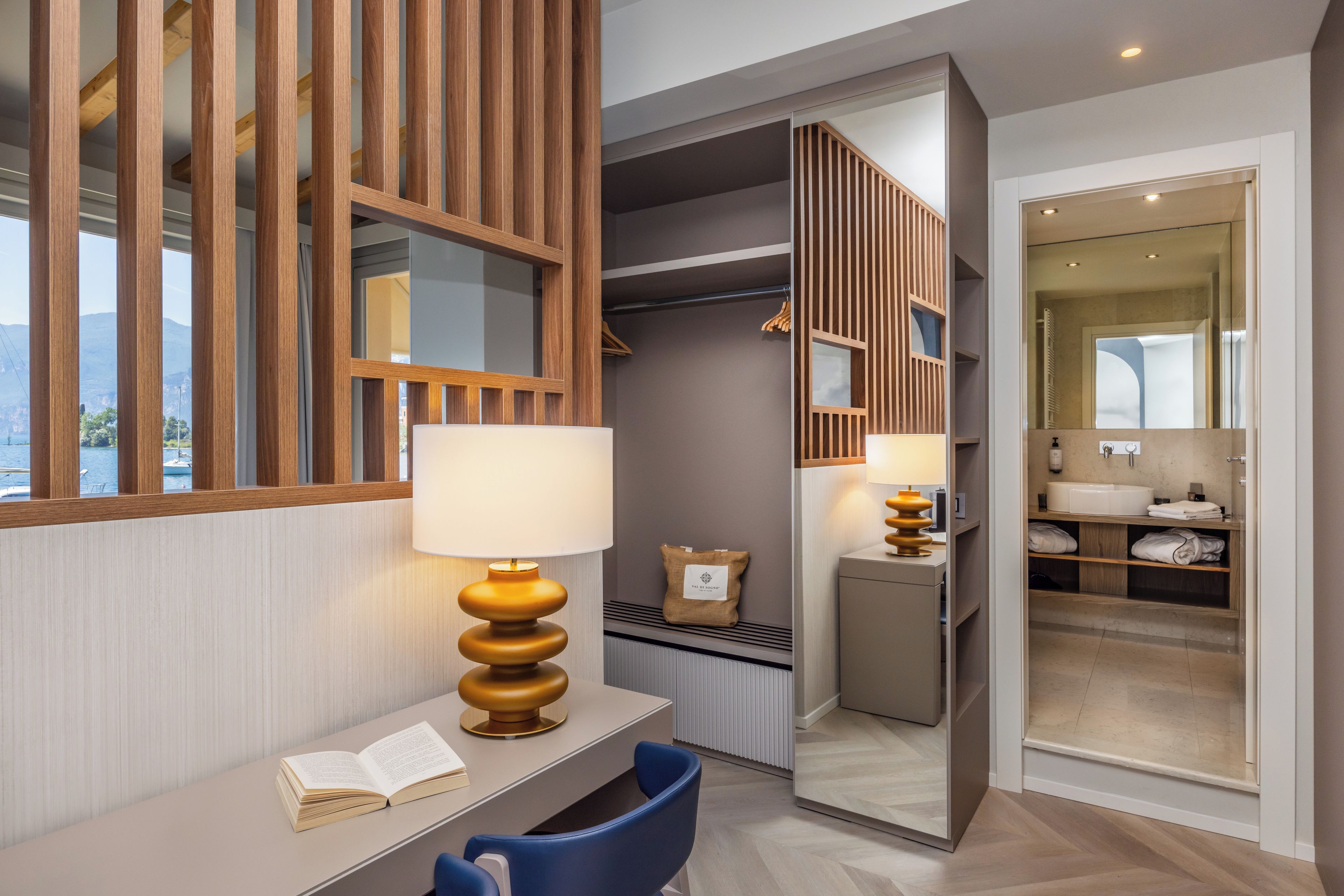 Una habitación de hotel moderna con un escritorio de madera, una silla azul, un espejo grande y un baño visible a través de una puerta abierta.