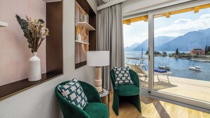 A modern hotel room with a large window overlooking a lake and mountains. There are two green armchairs with patterned cushions, a white vase with dried flowers, and a lamp on a side table. - Hotel Val di Sogno (Malcesine)