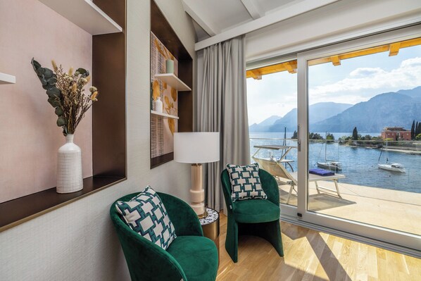 A modern hotel room with a large window overlooking a lake and mountains. There are two green armchairs with patterned cushions, a white vase with dried flowers, and a lamp on a side table. - Hotel Val di Sogno (Malcesine)