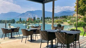 An outdoor seating area with tables and chairs, overlooking a lake with boats and a mountainous backdrop. - Hotel Val di Sogno (Malcesine)